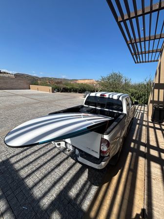 White pickup truck parked under a slatted pergola casting striped shadows, longboard surfboard sticking out of the truck bed against a clear blue sky and arid hills.