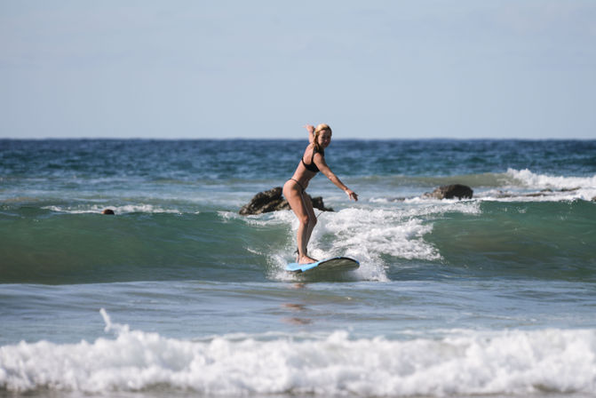 Female surfer balancing on a blue longboard, riding a small green wave toward the sandy shore with rocky outcrops and open ocean in the background