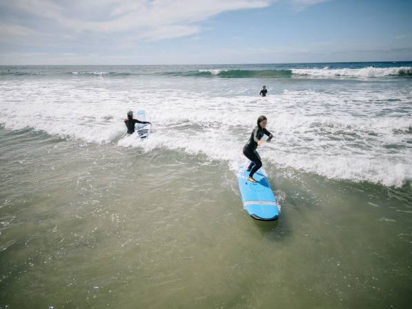 Group of surfers in wetsuits riding small waves on blue surfboards near a sandy ocean beach under a bright, partly cloudy sky.