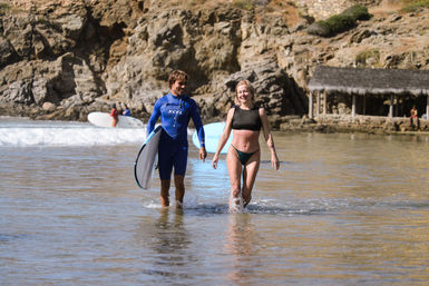 Two surfers emerging from shallow water at a sunny rocky cove — man in a blue wetsuit carrying a surfboard and woman in a bikini splashing water, cliffs and a beach hut in the background.