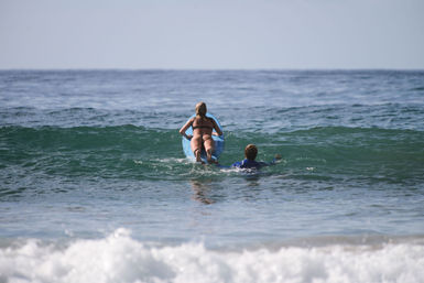 Adult and child paddling out on a blue surfboard through a small green wave at the beach, seen from behind — sunny ocean surf outing.