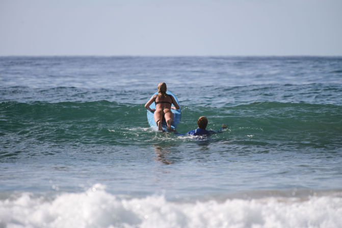 Adult and child paddling out on a blue surfboard through a small green wave at the beach, seen from behind — sunny ocean surf outing.
