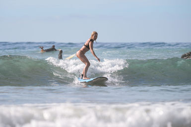 Woman in a black bikini surfing a small ocean wave on a blue board with other surfers paddling in the background at a sunny beach