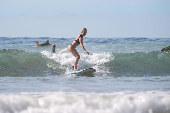 Woman in a black bikini surfing a small ocean wave on a blue board with other surfers paddling in the background at a sunny beach