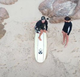Aerial view of two people on a sandy beach by rocks — one kneeling on a white longboard surfboard while the other watches, a longboard surf lesson on a rocky shoreline.