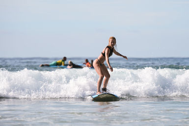 Woman balancing on a blue surfboard riding a small breaking wave near shore, ankle leash visible and other surfers paddling in the background on a sunny ocean day