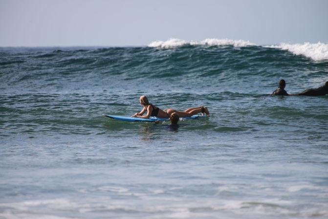 Surfer paddling on a blue surfboard with a companion nearby in the sunlit ocean, heading toward rolling waves