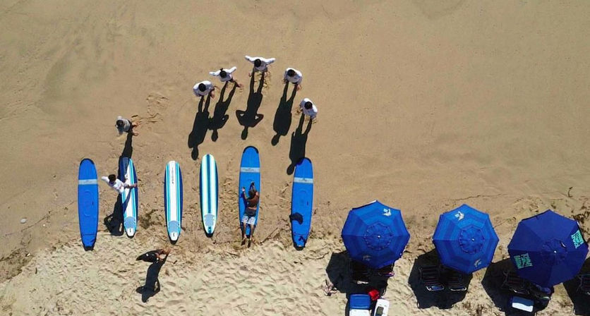 Aerial drone view of a sandy beach with six people standing near a row of blue and white surfboards and three blue beach umbrellas