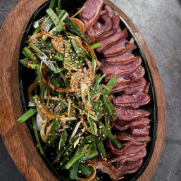 Sizzling cast-iron platter with thinly sliced seared meat alongside a scallion-and-onion salad tossed with sesame seeds and chili glaze, served on a wooden board.
