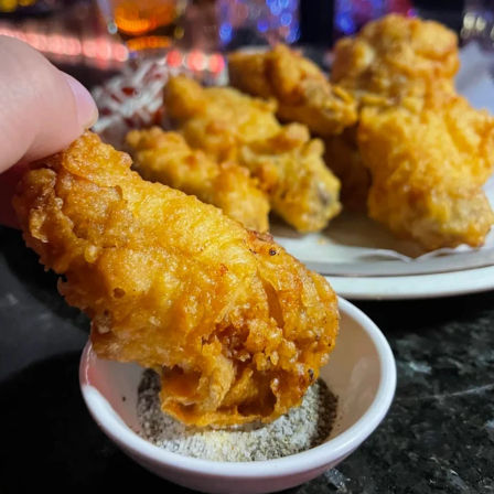 Crispy golden fried chicken wing held over a small bowl of peppery dipping salt, more wings piled on a plate on a dim bar counter