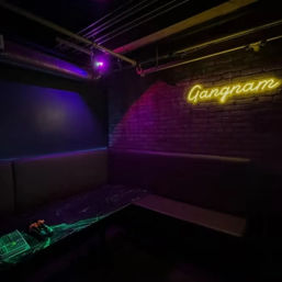 Dim neon-lit karaoke booth with brick wall and yellow script neon, purple and green spotlights, cushioned corner seating and a low table holding a microphone and control panel