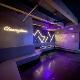 Modern purple-lit karaoke lounge interior with wraparound booth seating, geometric LED wall accents, neon script sign and low tables reflecting on a glossy floor.