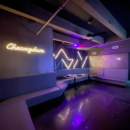 Modern purple-lit karaoke lounge interior with wraparound booth seating, geometric LED wall accents, neon script sign and low tables reflecting on a glossy floor.