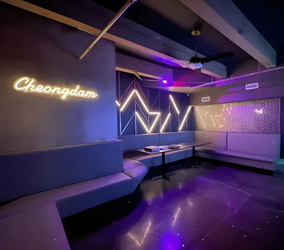 Modern purple-lit karaoke lounge interior with wraparound booth seating, geometric LED wall accents, neon script sign and low tables reflecting on a glossy floor.