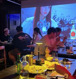Group dining in a cozy, dim Korean-style bar with shared plates, soju bottles and a small table lamp, colorful animated music video projected across the wall