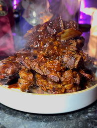 Close-up of a steaming stack of sticky, charred barbecue ribs glazed in smoky reddish-brown sauce on a white plate — a mouthwatering BBQ platter.