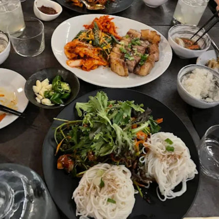 Korean restaurant table spread with grilled pork belly and spicy kimchi on a white plate, bowls of rice and banchan, a small dish of garlic and green chili, and a black plate of fresh greens with two nests of white somyeon noodles.