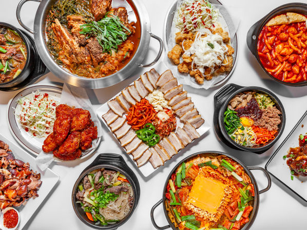 Top-down view of a colorful Korean food feast featuring spicy tteokbokki, cheese-topped ramyeon stew, bibimbap, sliced pork belly, kimchi stew, crispy Korean fried chicken and assorted side dishes on a white table.