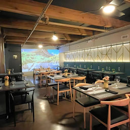 Cozy modern restaurant interior with exposed wooden ceiling beams, geometric backlit wall panels, rows of set tables and wooden chairs with wine glasses and small flower vases, and a large projected outdoor mural adding a relaxed, ambient vibe.