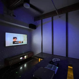 Private karaoke room with wall-mounted TV showing subtitles, moody purple LED lighting, low black table holding microphones and a remote, bench seating and ceiling fan.