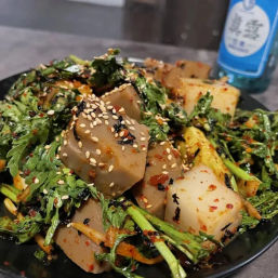 Close-up of spicy Korean acorn-jelly salad (dotori-muk) in a black bowl, tossed with fresh greens, chili flakes and toasted sesame seeds.