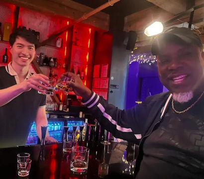 Two people toasting at a neon-lit cocktail bar — smiling bartender and patron clinking whiskey glasses over a glossy black counter with backlit bottles and festive lights.