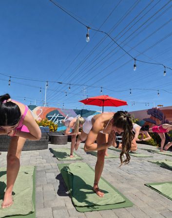 Sunlit rooftop yoga class with people balancing on one leg on green mats under string lights and a red umbrella, colorful mural wall and clear blue sky in the background.