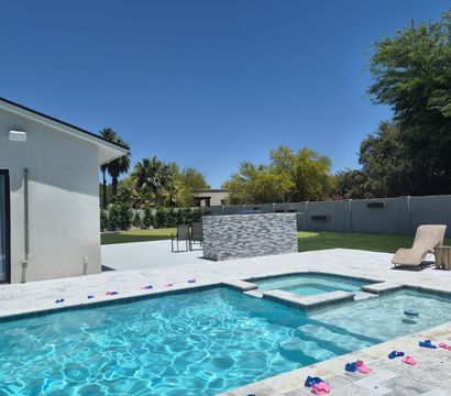 Modern backyard with turquoise swimming pool and attached spa, marble-look tile deck dotted with pink and blue pool toys, wicker lounge chair and stacked-stone outdoor bar, palm trees under a clear blue sky.