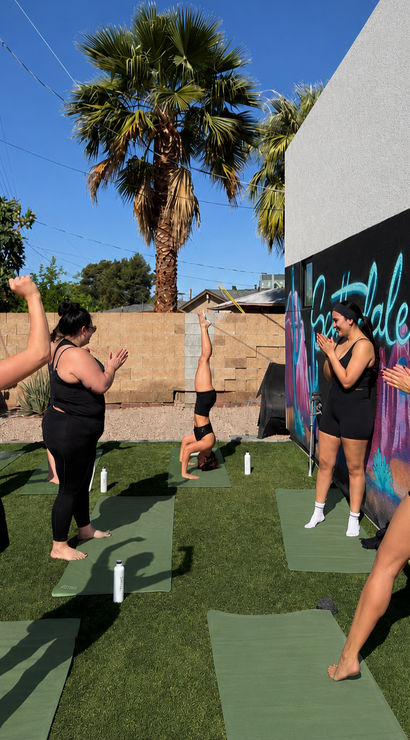 Outdoor yoga class on green mats in a sunny backyard with palm trees and a colorful mural — women in black activewear cheer as one woman holds a headstand.