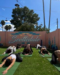 Group practicing outdoor yoga in child's pose on green mats in front of a pastel pink Scottsdale mural with cacti, string lights and palm trees under a sunny blue sky.