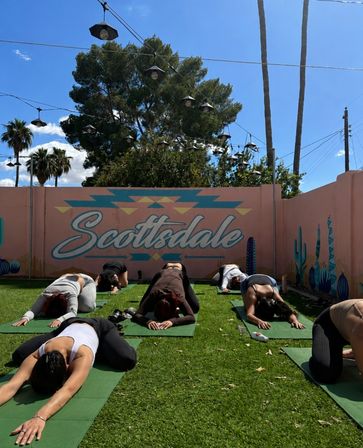 Group practicing outdoor yoga in child's pose on green mats in front of a pastel pink Scottsdale mural with cacti, string lights and palm trees under a sunny blue sky.