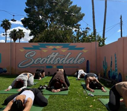 Group practicing outdoor yoga in child's pose on green mats in front of a pastel pink Scottsdale mural with cacti, string lights and palm trees under a sunny blue sky.