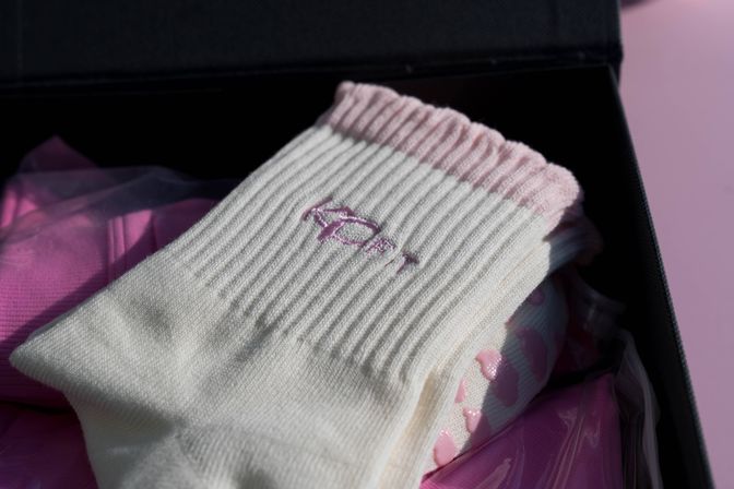 White ribbed socks with a pink cuff and small pink embroidered detail, pink non-slip dots visible, presented in a black gift box on a soft pink background.