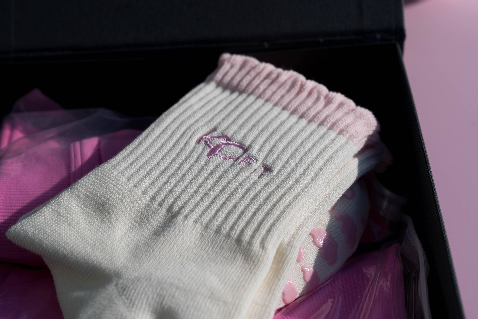 White ribbed socks with a pink cuff and small pink embroidered detail, pink non-slip dots visible, presented in a black gift box on a soft pink background.