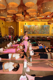 Group yoga class in a warm indoor studio — instructor in pink guiding participants in savasana on pink mats beneath large woven pendant lights and a colorful mural