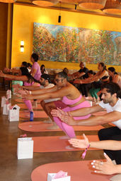 Energetic group fitness class in a bright indoor studio, participants in pink activewear doing squats on matching mats beneath a large colorful mural and warm yellow lighting.
