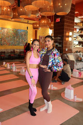 Two smiling women in bright activewear pose on pink yoga mats in a boutique fitness studio set up for a women's yoga event, with woven pendant lights, a colorful wall mural and gift bags in the background.