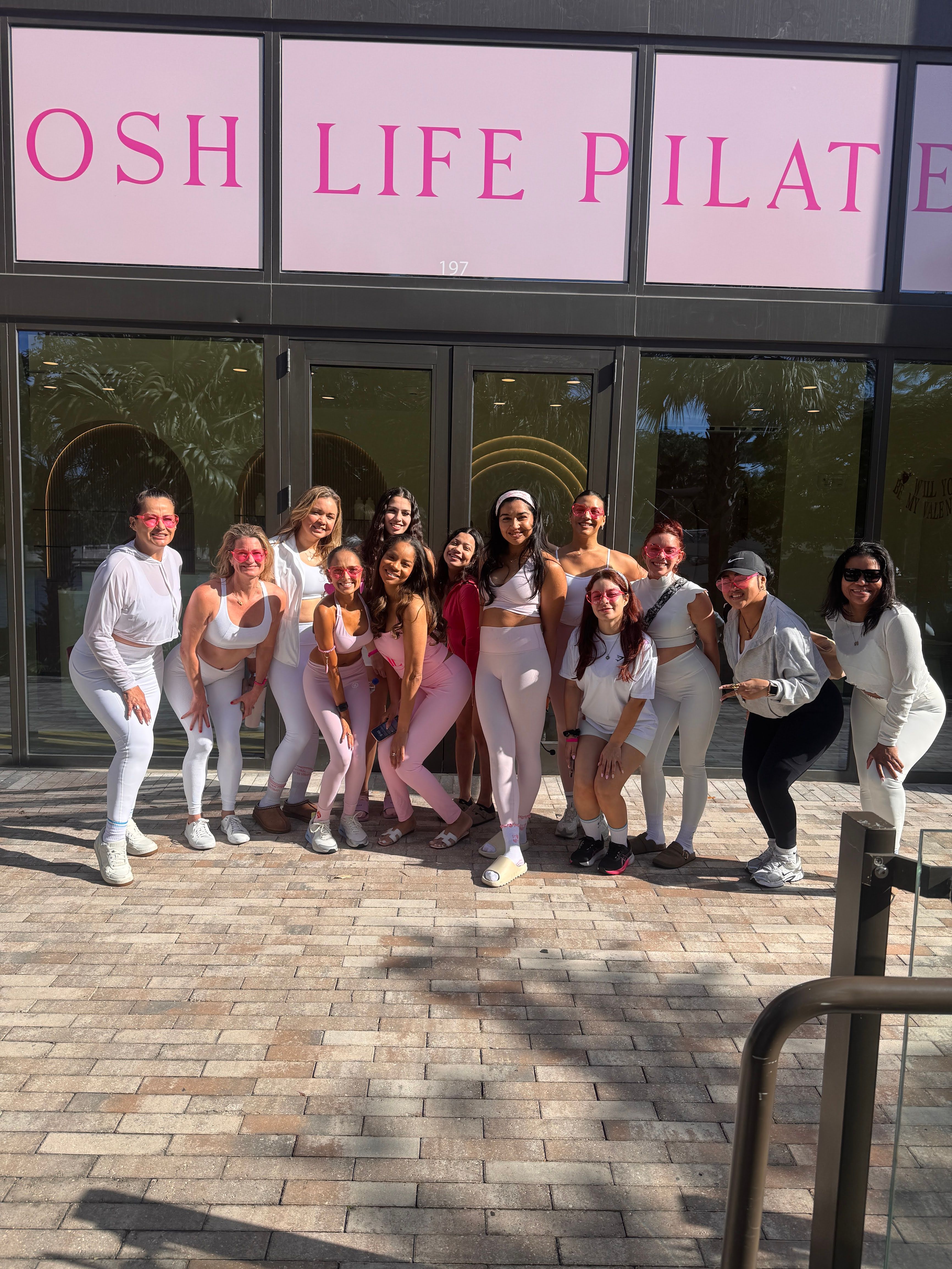 Smiling group of women and girls in pink and white activewear posing outside a pilates studio with pink signage on a sunny sidewalk.