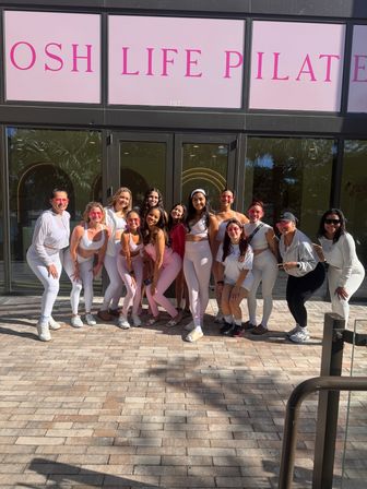 Smiling group of women and girls in pink and white activewear posing outside a pilates studio with pink signage on a sunny sidewalk.