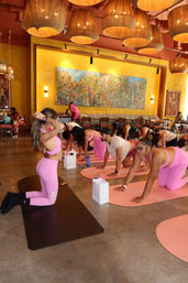 Group indoor yoga class in a bright boho studio — participants on pink mats and matching activewear practicing hands-and-knees poses under large woven pendant lights in front of a colorful mural.