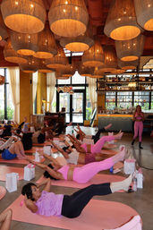 Group core workout in a boutique urban studio with oversized woven pendant lights and a bar in the background, participants on pink mats wearing colorful activewear and an instructor leading the class.