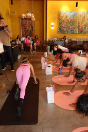 Community indoor yoga class in a warm yellow studio — participants in pink activewear on mats doing plank poses, with gift bags and a photographer.
