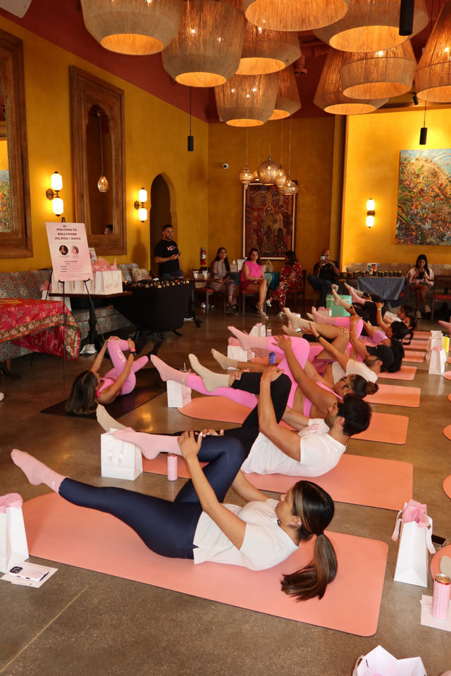 Group pilates class in a warm, Moroccan-inspired boutique studio: participants on pink mats doing leg-hold exercises under woven pendant lights, with gift bags and seated guests in the background.