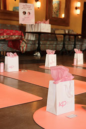 Pink yoga mats and white gift bags with pink tissue paper neatly arranged on a polished studio floor for a pilates/wellness class in a cozy lounge setting.