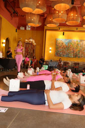 Group yoga relaxation class in a bright indoor studio — participants lying on pink mats while an instructor in pink leads the session under large woven pendant lamps and warm yellow walls with vibrant artwork.