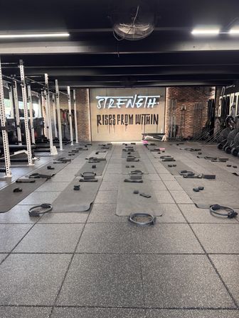Industrial-style fitness studio, rows of black exercise mats with Pilates rings and small weights set for class, brick back wall mural reading STRENGTH RISES FROM WITHIN, squat racks and barbells to the left.