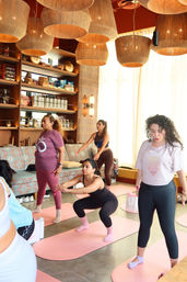 Women practicing yoga and squats on pink mats in a cozy sunlit studio with large woven pendant lights, patterned seating and shelves of decorative items.