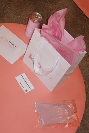 Playful pink-themed gift setup with a white paper bag stuffed with pink tissue, a pink canned drink, an envelope and business card, and a packaged pink garment arranged on a coral round table.