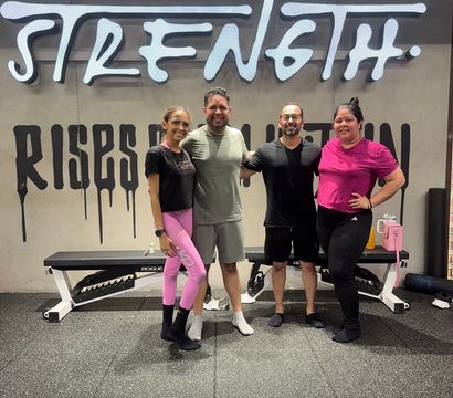 Smiling group of four adults in workout clothes posing arm-in-arm in a gym in front of a large 'STRENGTH' wall mural, with a bench and rubber gym flooring visible.