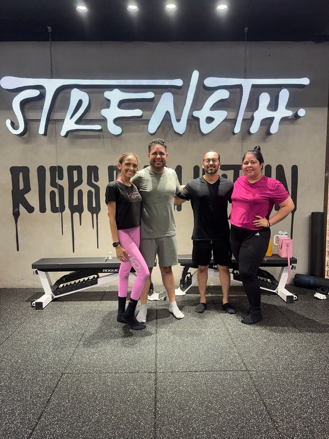 Smiling group of four adults in workout clothes posing arm-in-arm in a gym in front of a large 'STRENGTH' wall mural, with a bench and rubber gym flooring visible.